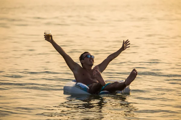 Gelukkig jongeman met opgeheven handen in de oceaan of de zee water met glas bier schreeuwen en geniet van de zomer roeping op op opblaasbare ring op zonsondergang — Stockfoto