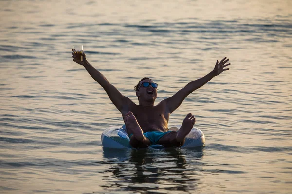 Молодой счастливый человек с поднятыми руками в океане или морской водой со стаканом пива кричит и наслаждаться летним призванием на надувном кольце на закате — стоковое фото