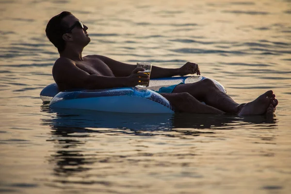 Молодой человек в солнечных очках отдыхает в океанской воде на надувном кольце на закате и пьет пиво. Летнее призвание — стоковое фото
