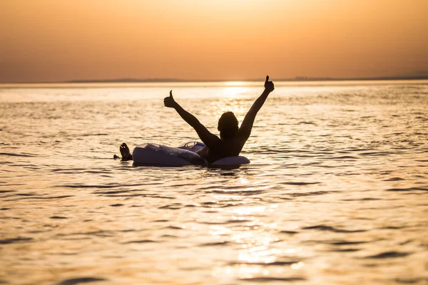 Gelukkig jongeman met opgeheven handen drijvend op opblaasbare ring in het water van de Oceaan op de zonsondergang. Man geniet van zomer roeping met handen omhoog in de oceaanwater. — Stockfoto