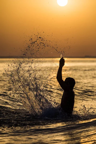 Hombre salpicando agua durante las vacaciones de verano al atardecer. Joven hombre atractivo divirtiéndose en una playa tropical al atardecer — Foto de Stock