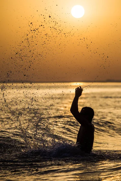 En mann som plasker vann i sommersolnedgangen. Ung, attraktiv mann som har det gøy på en tropisk strand ved solnedgang – stockfoto