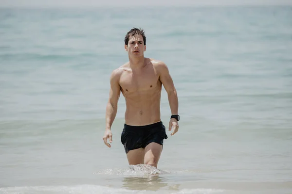 Young handsome muscular man walking out of the water in a tropical beach wearing a bathing suit — Stock Photo, Image