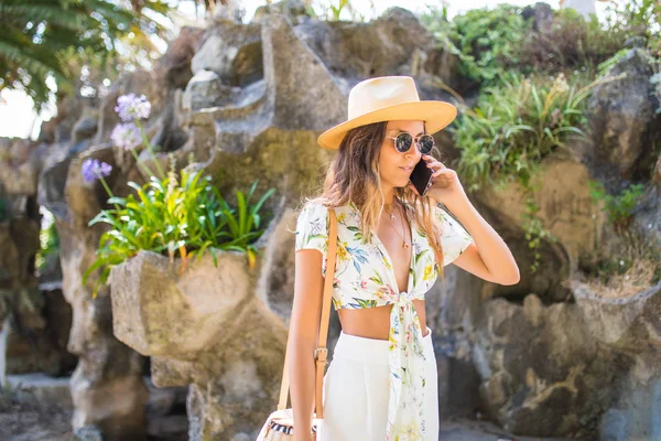 Menina sorridente atraente falando no telefone andando em um parque em chapéu e óculos de sol . — Fotografia de Stock