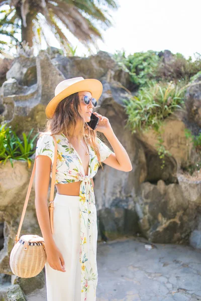 Menina sorridente atraente falando no telefone andando em um parque em chapéu e óculos de sol . — Fotografia de Stock