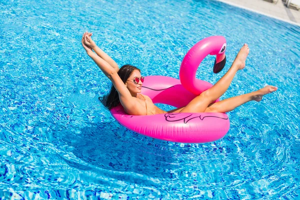 Femeie frumoasă, purtând costum de baie, întinsă pe o saltea de aer roz flamingo într-o piscină cu apă albastră, vară — Fotografie, imagine de stoc