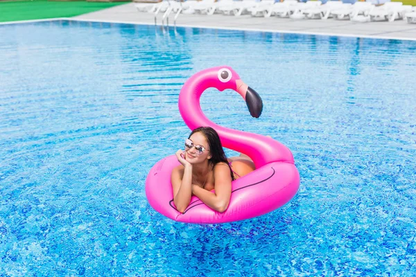 Fashion portrait of a young and sexy girl in the pool on an inflatable pink flamingo in a bathing suit and sunglasses in summer — Stock Photo, Image