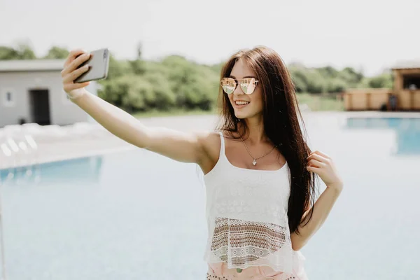 Close-up selfie-retrato de menina morena atraente com cabelos longos em pé perto da piscina. Ela usa t-shirt rosa, óculos de sol . — Fotografia de Stock
