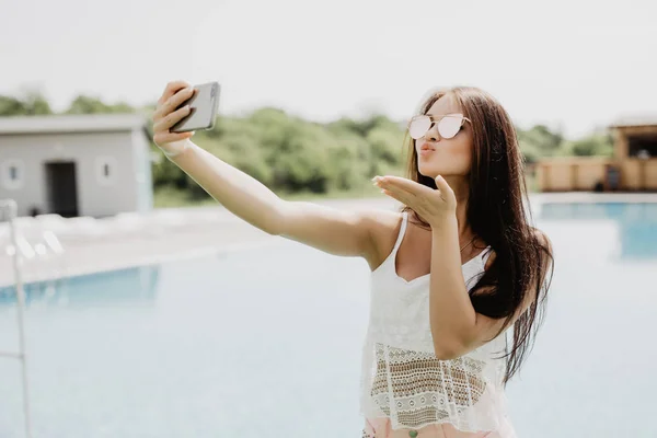 Close-up selfie-portrait of attractive brunette girl with long hair standing near pool. She wears pink T-shirt, sunglasses. — Stock Photo, Image