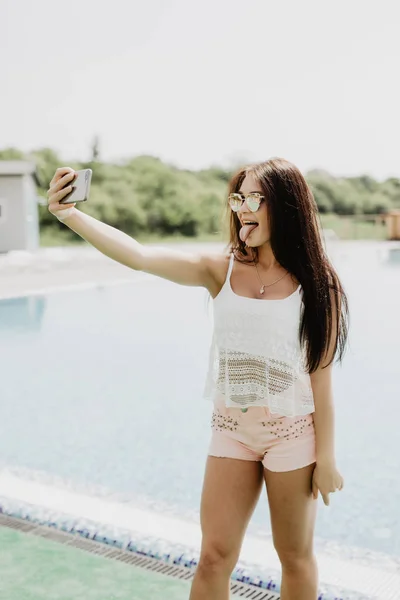 Närbild selfie-porträtt av attraktiv brunett tjej med långt hår står nära poolen. Hon bär rosa T-shirt, solglasögon. Hon ler till kameran och visar cool look. — Stockfoto