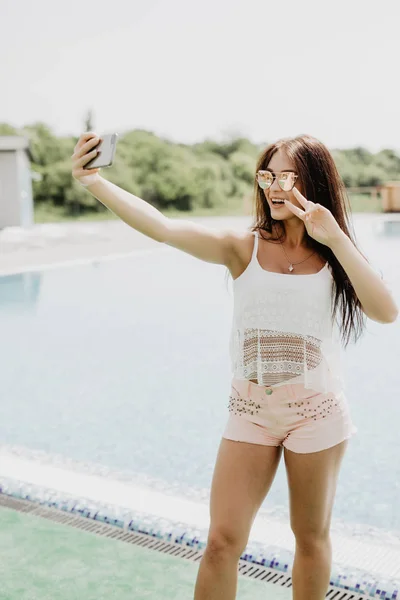 Close-up selfie-retrato de menina morena atraente com cabelos longos em pé perto da piscina. Ela usa t-shirt cor-de-rosa, óculos de sol. Ela está sorrindo para a câmera e mostra olhar legal . — Fotografia de Stock