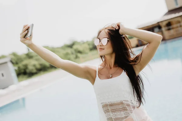 클로즈업 selfie-수영장 근처에 서 긴 머리와 매력적인 갈색 머리 여자의 초상화. 그녀는 분홍색 t-셔츠, 선글라스를 착용. 그녀는 카메라에 웃 고 멋진 모습을 보여줍니다.. — 스톡 사진