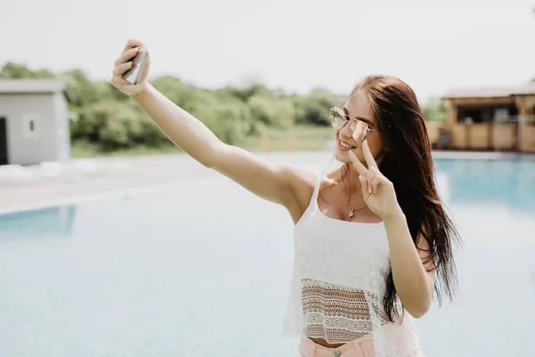 Primer plano selfie-retrato de chica morena atractiva con el pelo largo de pie cerca de la piscina. Lleva una camiseta rosa, gafas de sol. Ella sonríe a la cámara y muestra una mirada fresca . — Foto de Stock