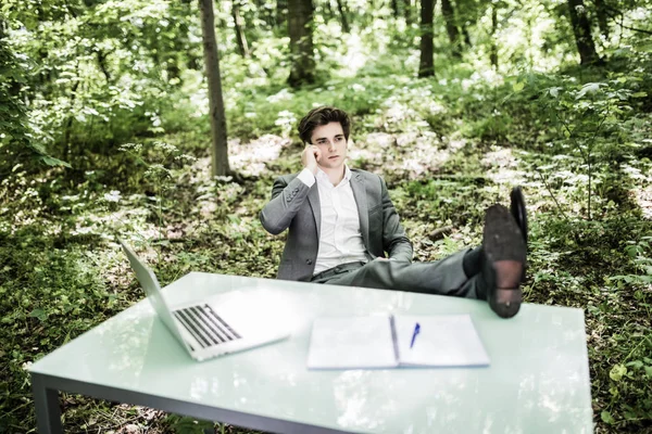 Business man talk on mobile phone at office table in green forest with legs on table. Business concept.