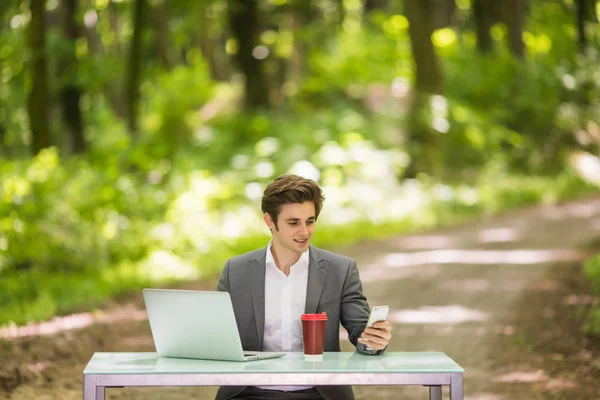 Ritratto di uomo d'affari seduto alla scrivania dell'ufficio con computer portatile e una tazza di caffè che parla telefono cellulare nel parco verde della foresta. Concetto aziendale . — Foto Stock