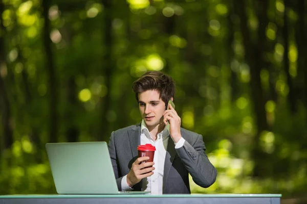 Retrato de Freelancer sentado en el escritorio de la oficina de trabajo en el ordenador portátil y una taza de café hablando de teléfono móvil en la carretera del parque forestal verde. Concepto empresarial . —  Fotos de Stock