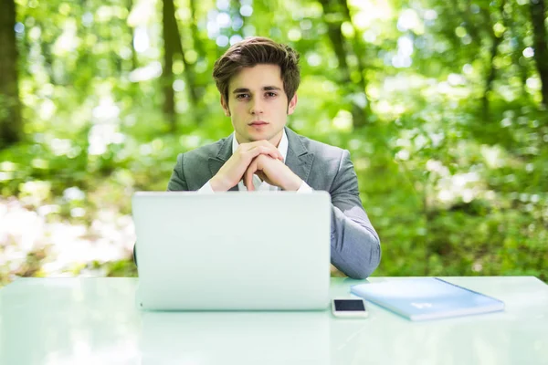 Il giovane uomo d'affari bello sopra computer portatile a tavolo di ufficio guarda la macchina fotografica in parco verde. Concetto aziendale . — Foto Stock