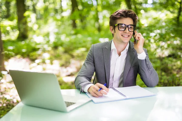 Ritratto di giovane bell'uomo d'affari che lavora al computer portatile al tavolo dell'ufficio e parla al telefono con il cliente nel parco verde della foresta. Concetto aziendale . — Foto Stock