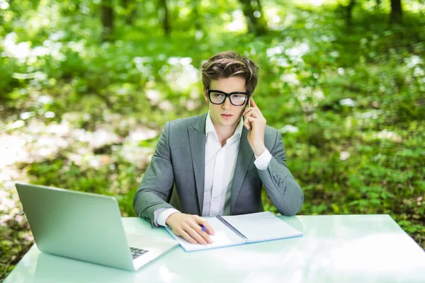 Ritratto di giovane affascinante manager che lavora al computer portatile al tavolo dell'ufficio e parla al telefono con il cliente nel parco verde della foresta. Concetto aziendale . — Foto Stock