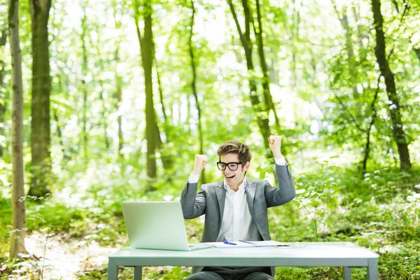 Ritratto di giovane bell'uomo d'affari in giacca e cravatta che lavora al computer portatile al tavolo dell'ufficio con le mani alzate obiettivi raggiunti nel parco verde della foresta. Concetto aziendale . — Foto Stock