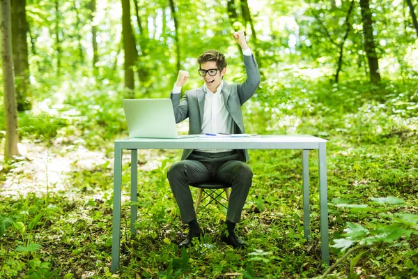Ritratto di giovane affascinante manager di successo in giacca e cravatta che lavora al computer portatile al tavolo dell'ufficio con le mani alzate nel parco verde della foresta. Concetto aziendale . — Foto Stock
