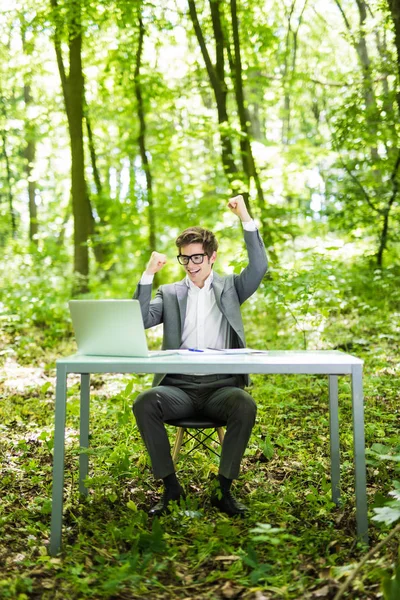 Ritratto di giovane affascinante freelance di successo in giacca e cravatta che lavora al computer portatile al tavolo dell'ufficio con le mani alzate nel parco verde della foresta. Concetto aziendale . — Foto Stock