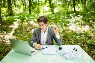 Genç yakışıklı iş adamı iş tablo ofisinde buruşuk kağıtlar duygu ile yeşil orman içinde laptop ile yorgun kızgın ve üzgün Hayır fikirler. İş kavramı