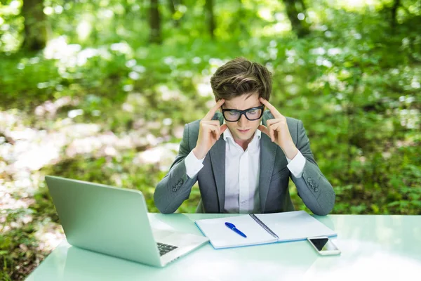Ritratto di giovane bell'uomo d'affari in giacca e cravatta che lavora al computer portatile al tavolo dell'ufficio con le mani sulla testa del dolore nel parco verde della foresta. Concetto aziendale . — Foto Stock