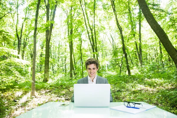 Ritratto di giovane bell'uomo d'affari in giacca e cravatta che lavora al computer portatile al tavolo dell'ufficio nel parco verde della foresta. Concetto aziendale . — Foto Stock