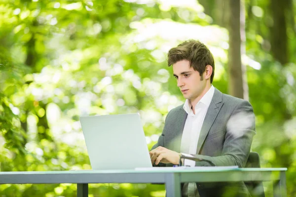 Ritratto laterale di giovane bell'uomo d'affari in giacca e cravatta che lavora al computer portatile al tavolo dell'ufficio nel parco verde della foresta. Concetto aziendale . — Foto Stock