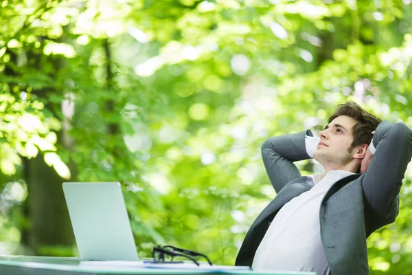 Ritratto di giovane bell'uomo d'affari in giacca e cravatta si rilassi a computer portatile a tavolo di ufficio passa sopra la testa in parco di foresta verde. Concetto aziendale . — Foto Stock