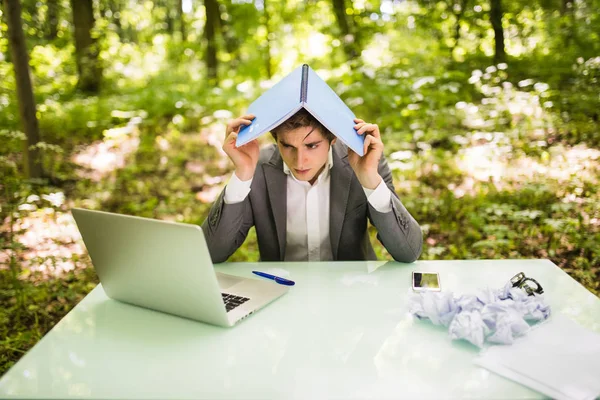 Młody przystojny biznesmen w urzędzie pracy tabela z laptopa w zielonym lesie z notebook nad głową, zbyt wiele pracy. Koncepcja biznesowa — Zdjęcie stockowe
