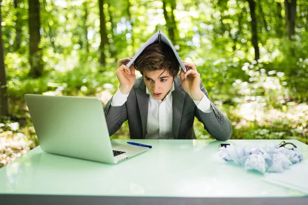 Giovane bell'uomo d'affari al tavolo da lavoro ufficio con computer portatile nella foresta verde con notebook sopra la testa, troppo lavoro. Concetto aziendale — Foto Stock