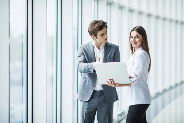 Empresario y mujer de negocios usando un ordenador portátil juntos mientras están de pie frente a las ventanas del edificio de oficinas con vistas a la ciudad — Foto de Stock