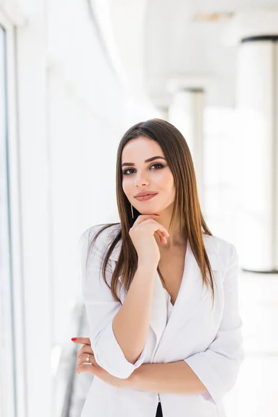 Portret van vertrouwen jonge zakenvrouw met gekruiste armen leunend tegen venster in kantoor — Stockfoto