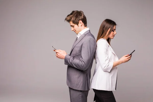 Casal jovem está usando telefones inteligentes e sorrindo enquanto está de costas para trás em um fundo cinza . — Fotografia de Stock