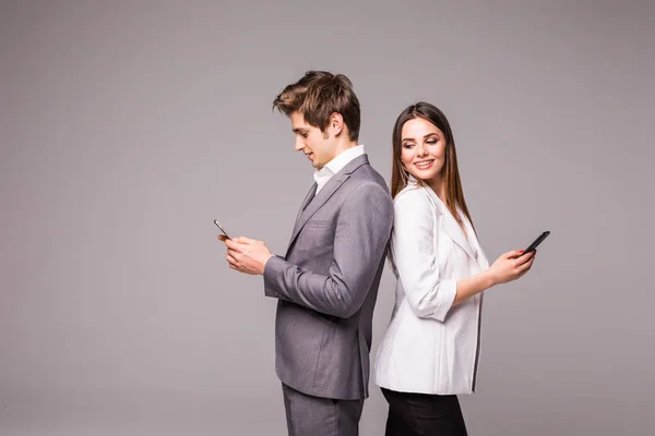 Casal jovem está usando telefones inteligentes e sorrindo enquanto está de costas para trás em um fundo cinza . — Fotografia de Stock