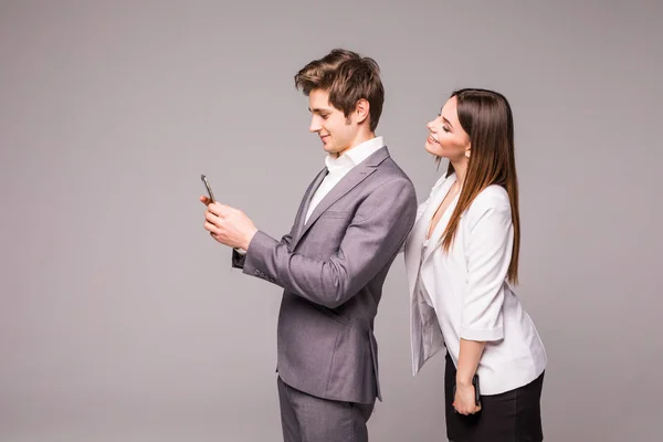 O jovem casal está usando telefones inteligentes e sorrindo enquanto está de costas para trás em um fundo cinza. Mulher olha para o homem . — Fotografia de Stock
