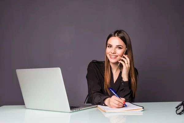 Buena charla de negocios. Joven alegre hermosa mujer en gafas hablando en el teléfono móvil y el uso de la computadora portátil con sonrisa mientras está sentada en su lugar de trabajo — Foto de Stock