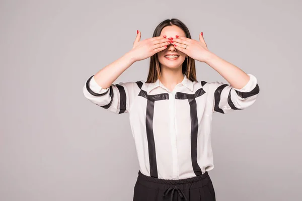 Portrait of young scared woman covering eyes with hands while standing against gray studio background. Confused girl close eyes with palms ignoring something