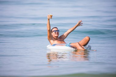 Eller yukarı bir bardak bira günbatımında kauçuk halka okyanus su üzerinde yüzen ile güneş gözlüğü ile genç adam
