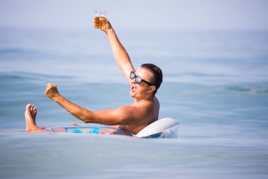 Bir bardak bira günbatımında kauçuk halka okyanus su üzerinde yüzen ile eller yukarı güneş gözlüğü ile genç adam alkış