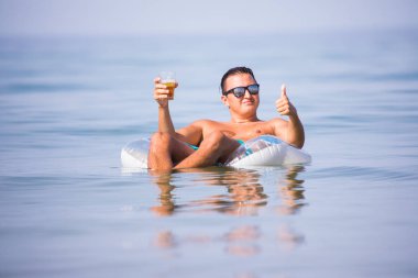 Başparmak yukarıya ile bir bardak bira kauçuk halka okyanus su üzerinde yüzen günbatımında güneş gözlüğü ile genç adam