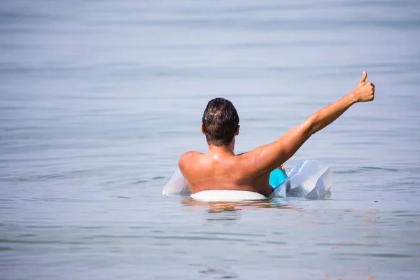 Joven hombre guapo con un vaso de cerveza sentado en círculo de flotador, disco de goma en el agua de mar con los pulgares hacia arriba . — Foto de Stock