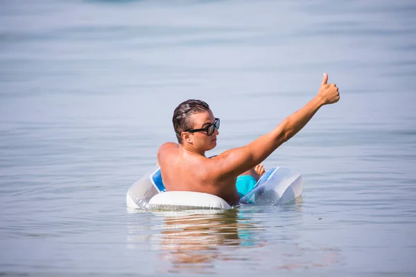 Jonge knappe man met een glas bier zit op float cirkel, rubber schijf in het water van de zee met duimen. — Stockfoto