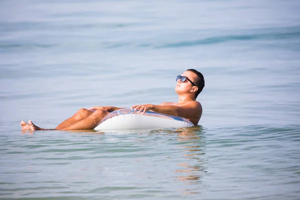 Молодой человек в солнечных очках со стаканом пива, плавающим на резиновом кольце в океанской воде — стоковое фото