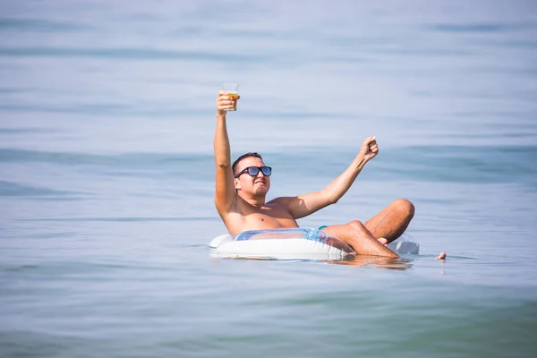 Joven con las manos en alto en gafas de sol con un vaso de cerveza flotando en el anillo de goma en el agua del océano al atardecer — Foto de Stock