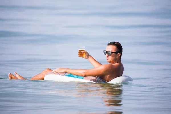 Молодой человек в солнечных очках с бокалом пива, плавающего на резиновом кольце в океанской воде на закате — стоковое фото