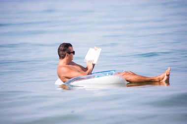 Genç adam sakin ol şişme halka bir kitap okumak okyanus su içinde. Yaz meslek