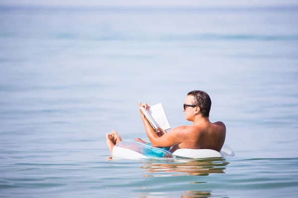 Молодой человек расслабляется в океанской воде на надувном кольце читает книгу. Летнее призвание — стоковое фото